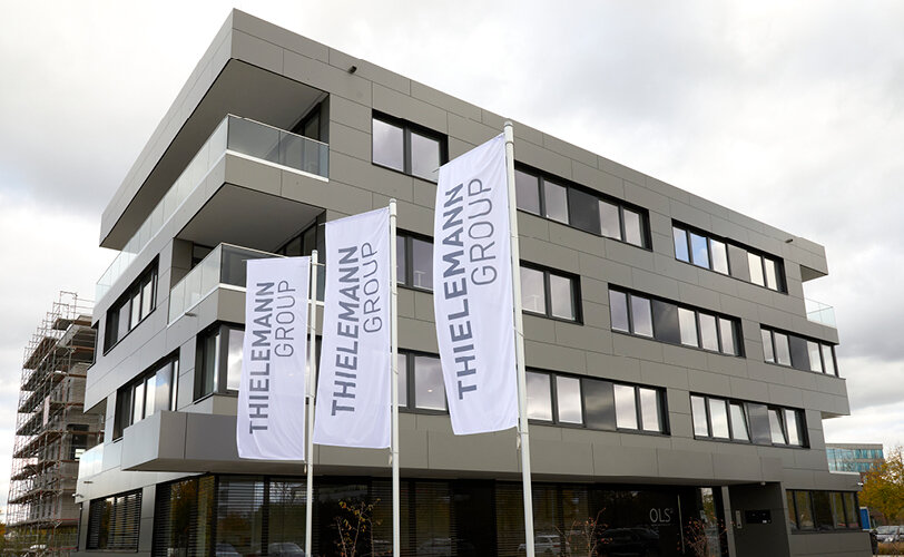 Foto: Graues Bürogebäude im Hintergrund, Fahnen im Vordergrund mit der Aufschrift und dem Logo der ThielemannGroup 
